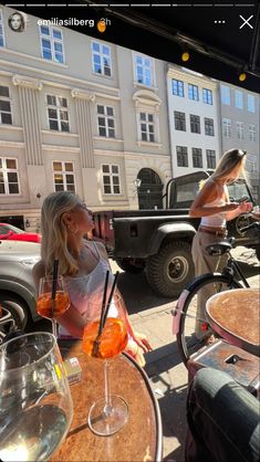 two women sitting at an outdoor table with drinks in front of some parked cars and buildings