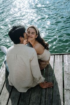 a man and woman are sitting on a dock by the water, one is kissing the other's cheek