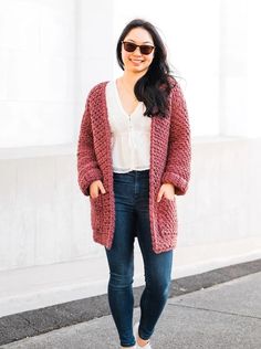 a woman standing in front of a building wearing a red cardigan sweater and jeans