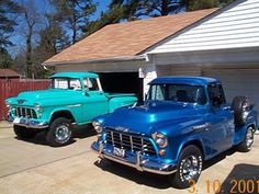 two old trucks parked in front of a house with garage doors open and one is blue