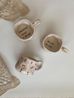 three coffee mugs with writing on them are sitting next to doily and lace