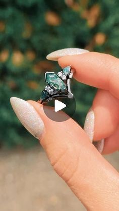 a woman's hand holding a ring with green and white stones on it, in front of some bushes