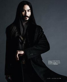 a man with long hair wearing a black coat and standing in front of a gray background