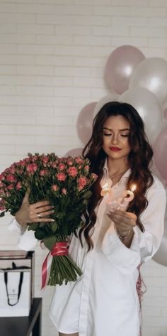 a woman holding flowers and candles in her hands with balloons behind her, on a white brick wall