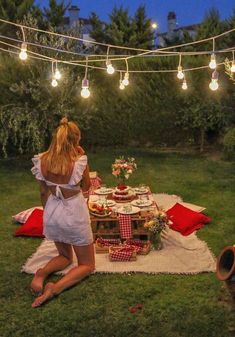 a woman sitting on the grass in front of a picnic table with food and drinks