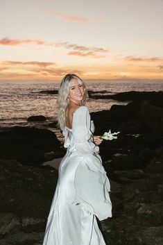 a woman in a long white dress standing on rocks near the ocean with her back to the camera