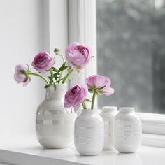 three white vases with pink flowers in them on a window sill next to a window