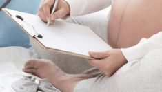 a pregnant woman writing on a clipboard while laying in bed with her stomach exposed