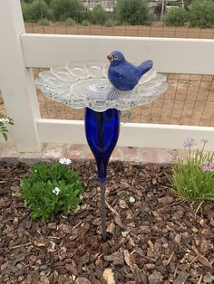 a blue glass bird feeder sitting on top of a flower bed next to a fence