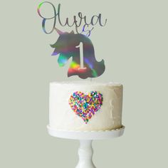 a white cake topped with a unicorn topper and rainbow sprinkles in the shape of a heart