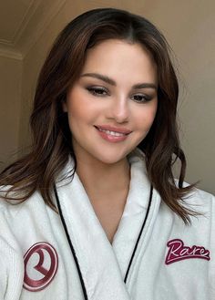 a woman with long hair wearing a white robe and smiling at the camera while standing next to a wall