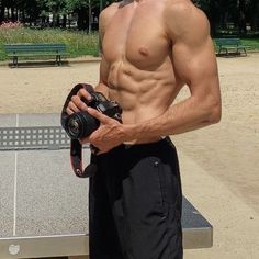 a shirtless man holding a camera in front of a table with benches behind him