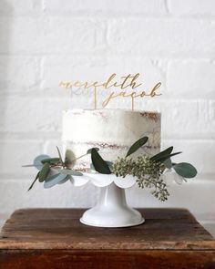 a white cake topped with greenery on top of a wooden table next to a brick wall