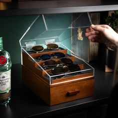 a person holding their hand over an open wooden box with sunglasses in it next to a bottle of alcohol