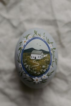 an egg painted with the image of a farm and white flowers is on a piece of paper