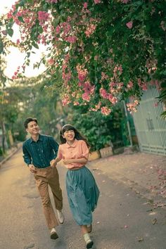 a man and woman walking down a street next to trees with pink flowers on them