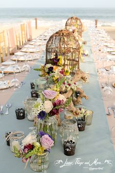 a long table with vases filled with flowers and candles on it next to the ocean