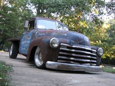 an old, rusty truck is parked on the side of a road in front of some trees