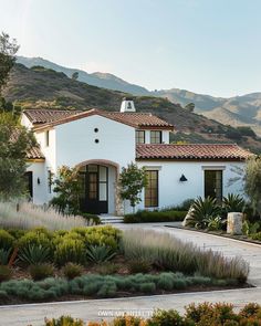 a white house with lots of plants and trees in front of the house, surrounded by mountains