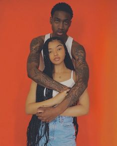 a man and woman standing next to each other in front of an orange wall with tattoos on their arms