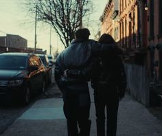 two people walking down the sidewalk next to parked cars