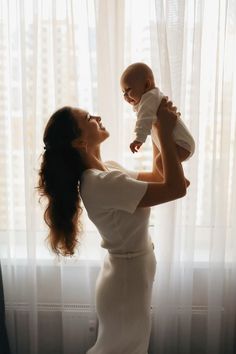 a woman holding a baby up to her face in front of a window with sheer curtains