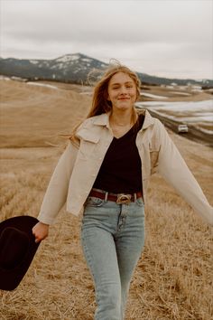 a woman walking through a field with a hat on her head and jacket over her shoulders