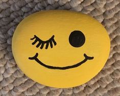 a smiley face painted on a yellow rock with black eyes and eyelashes, sitting on a woven surface