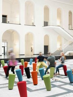 several people are walking around in a large building with many colorful stools on the floor