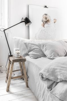 a bed with white sheets and pillows next to a wooden stool in front of a window