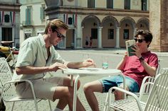 two young men sitting at a table in the middle of an open city square, one reading a book