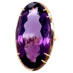 This large enchanting 14.50 carat faceted elongated oval amethyst is set in a mid-century modern 14 karat yellow gold setting that has a rose gold hue. This amethyst's rich deep purple comes alive in the light enhancing its brightness. The stone measures approximately 1 1/16" long by 1/2" wide and is held in the mounting by four stations of double prongs. Under the gem, the mounting appears as a frame highlighting the beauty of the stone in this circa 1970 mounting making the top of the ring approximately 1 1/4" long by 5/8" wide. This vibrant amethyst is very clean with no visible inclusions. The ring rises approximately 7/16" off the finger, is a US size 6.5, and weighs 9.1 grams. The 1970's was a decade of punk rock and disco, social change, and iconic movies such as The Godfather, Star Luxury Formal Rings With Oval Cabochon, Luxury Formal Amethyst Ring Oval Cabochon, Luxury Hallmarked Amethyst Oval Cabochon Ring, Luxury Purple Oval Cabochon Rings, Luxury Hallmarked Oval Cabochon Amethyst Ring, Luxury Oval Amethyst Ring In Art Deco Style, Luxury Oval Amethyst Ring, Art Deco Style, Luxury Yellow Gold Amethyst Cabochon Ring, Luxury Vintage Amethyst Oval Cabochon Ring