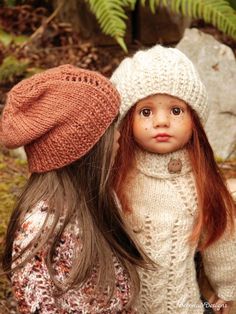 two dolls are standing next to each other in front of some trees and leaves, one is wearing a hat