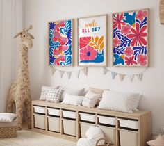 a white room with three colorful paintings on the wall and two stuffed giraffes