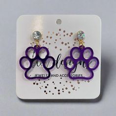 Acrylic Paw Earrings Show your team spirit with these fun paw-print earrings. The hollow acrylic paw design and iridescent gem stud create an eye-catching look. These lightweight earrings are perfect for cheering on your favorite team. Choose from black or purple to match your school colors. Details 1 1/4" overall length Stainless steel post earrings Iridescent gem stud Hollow acrylic paws Two colors, black or purple Handmade by a woman-owned business in Michigan Paw Earrings, Paw Print Earrings, Stationary Gifts, Paw Design, Steel Post, Lightweight Earrings, School Colors, Light Weight Earrings, Kids Jewelry