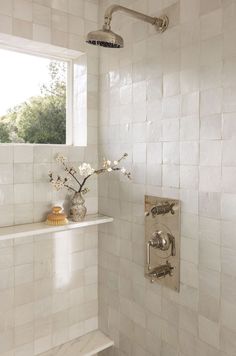 a white tiled bathroom with a window and shower head in the corner, next to a shelf with flowers on it