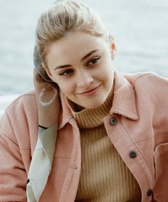 a woman with blonde hair wearing a pink jacket and white tie sitting next to the water