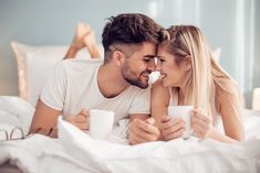 a man and woman laying in bed holding mugs looking into each others'eyes