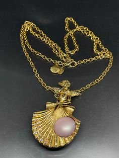 This vintage Kirks Folly necklace is a stunning piece of jewelry that will complement any outfit. The necklace features a gold-tone chain with a lobster closure and a pendant in the shape of a shell that also can be used as a brooch. The shell is adorned with Aurora Borealis crystals and a purple imitation pearl. The pendant also has a beautiful cherub or angel figure with crystal accents. This necklace is 30 inches long, making it the perfect length to wear with any neckline. It is a pre-owned vintage piece of jewelry that has been well-maintained and signed by the brand. Add this unique and glamorous necklace to your collection today! Evening Brooch Pendant Necklaces, Pendant Necklaces With Brooch For Evening, Elegant Gold Necklace With Brooch, Gold Pendant Necklace With Brooch, Vintage Necklace With Large Pendant For Evening, Vintage Evening Necklace With Large Pendant, Vintage Large Pendant Necklace For Evening, Vintage Gold-tone Necklace With Large Pendant, Angel Cherub