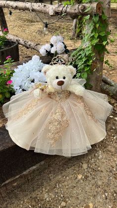 a teddy bear with a tiara sitting on a bench next to flowers and trees