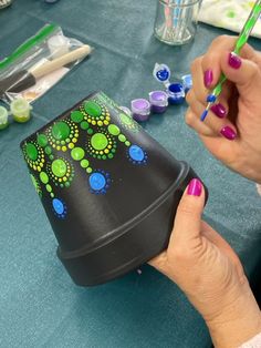 a woman is painting on a black vase with green and blue dots, while another person holds a pencil in their left hand