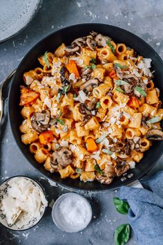 a skillet filled with pasta and mushrooms on top of a gray table next to two bowls