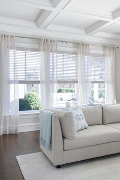 a living room filled with furniture and windows covered in white drapes on top of wooden flooring