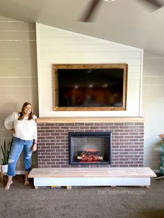 a woman standing next to a brick fireplace in a living room with a flat screen tv above it