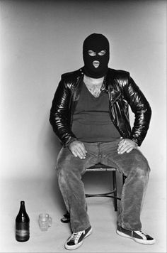 a man wearing a mask sitting on a chair next to a beer bottle and glass