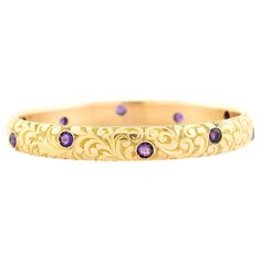 A beautiful gold and amethyst bangle bracelet from the Art Nouveau (ca1900) period! This stunning 14kt gold bracelet features a carved scrolling design that carries around the entire outer surface of the bracelet, adding wonderful texture and depth. Decorating the outside of the bangle are ten round cut amethysts, which rest in staggered bezel settings. The stones have a luscious natural purple color that looks fantastic paired with the vibrant gold setting. The slip-on style bangle has a smooth Amethyst Bangle, Amethyst Stone, Gold Design, 14kt Gold, Bangle Bracelet, Purple Color, A Group, Bangle Bracelets, Art Nouveau