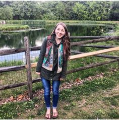 a woman standing next to a wooden fence near a lake with trees in the background