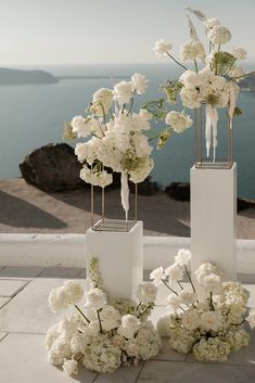 three white vases with flowers in them sitting on the ground next to water and rocks