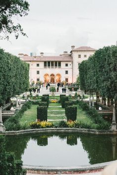 a large white building surrounded by lush green trees and bushes next to a small pond