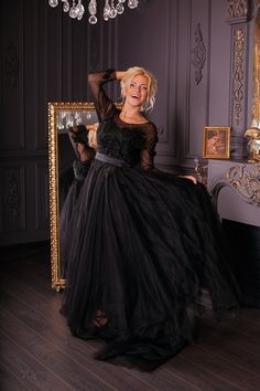 a woman in a long black dress posing for a photo with a mirror and chandelier